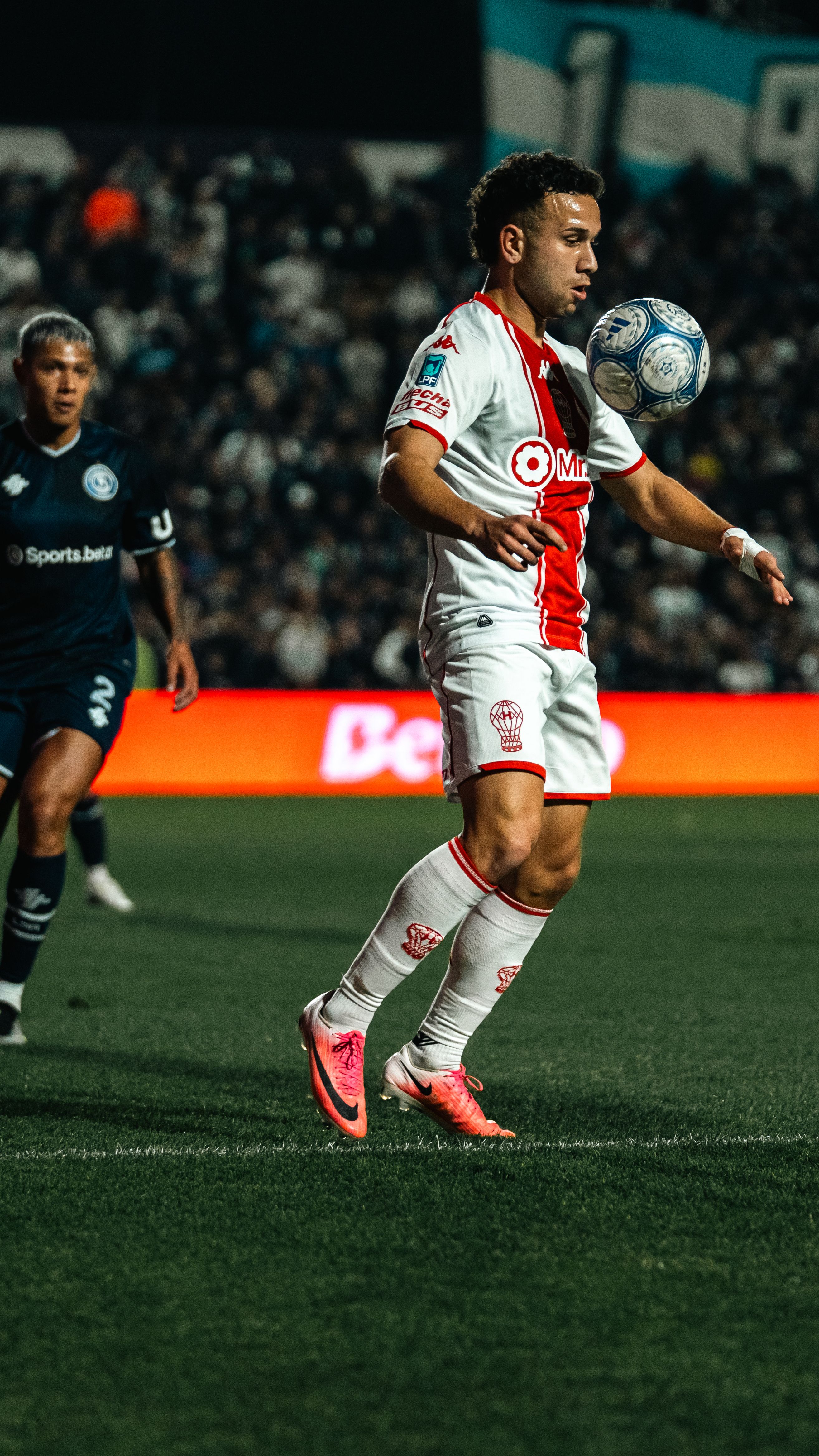 Soccer player in white and red kit balancing ball during an intense match at night.