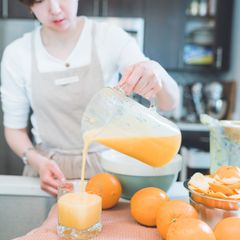 Woman in an apron pouring fresh orange juice from a jug into a glass, oranges on the kitchen worktop