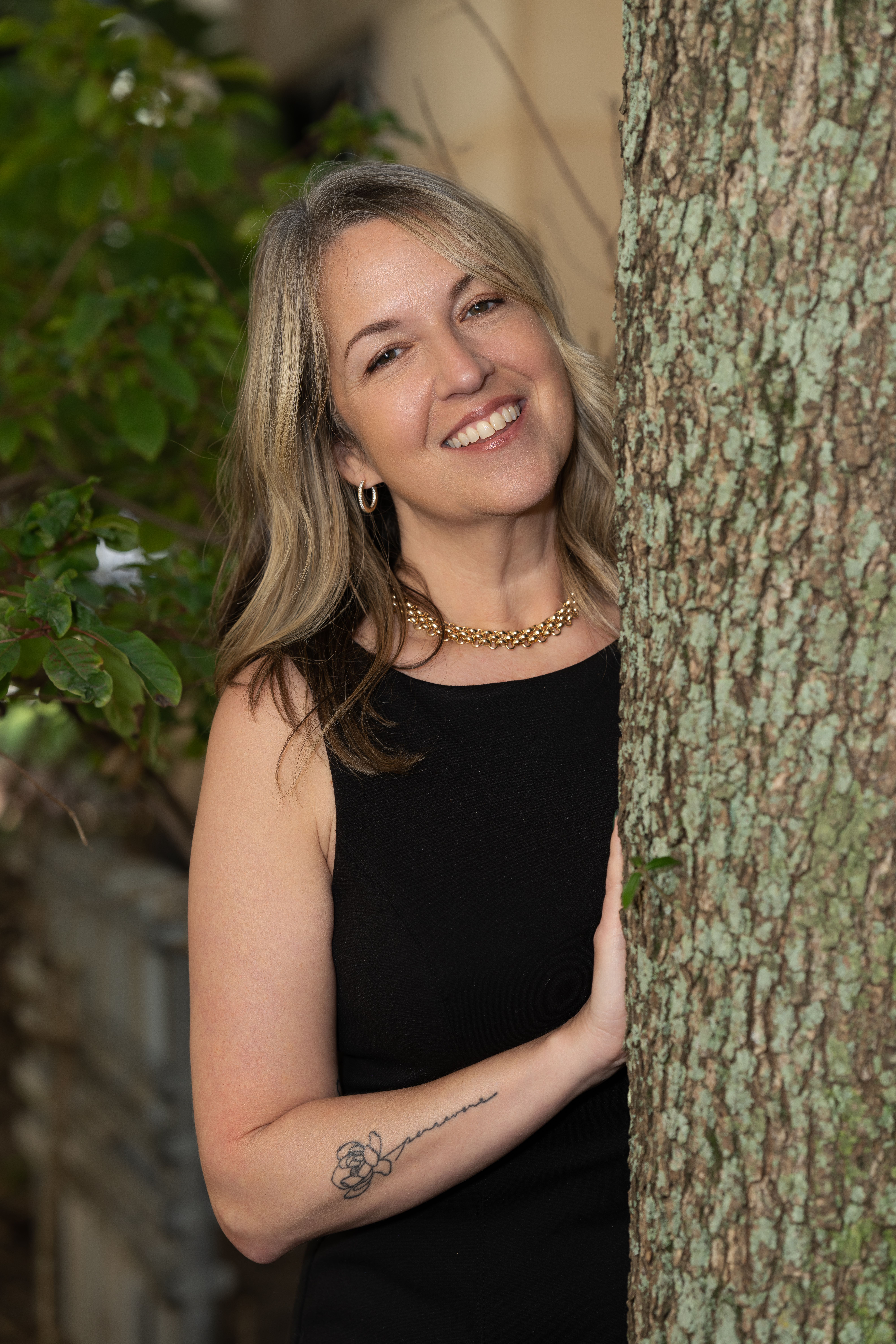 Smiling woman in elegant black dress standing outdoors by a tree trunk, exuding happiness.