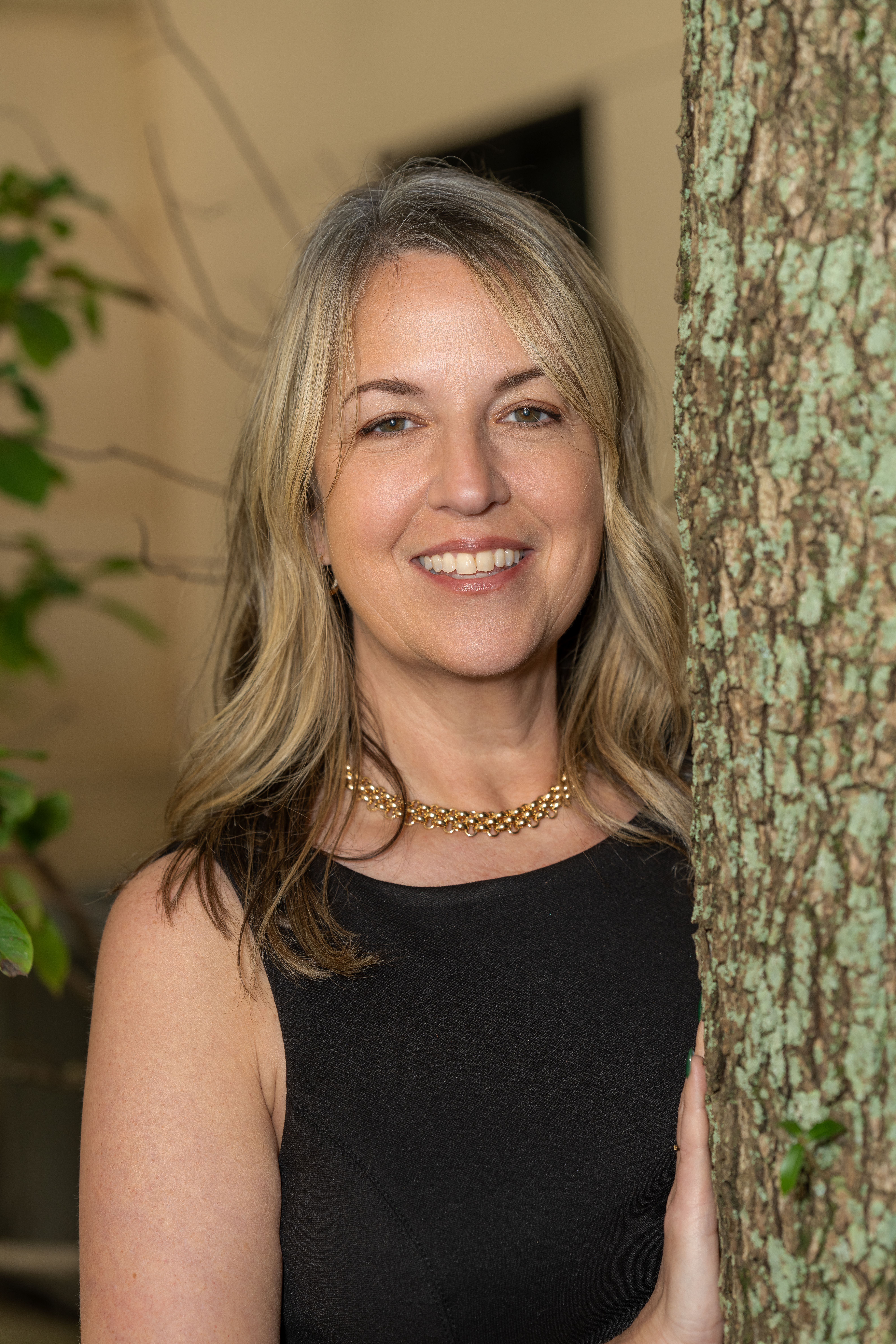 Blonde woman smiling warmly behind a tree outdoors.