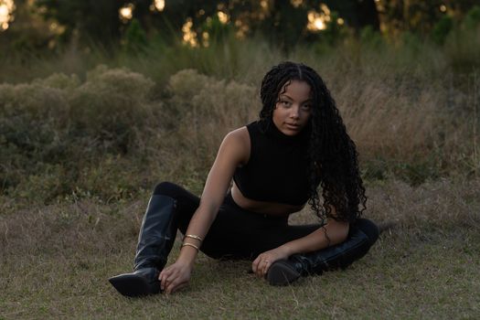 Young woman in black outfit sitting outdoors on grass, surrounded by nature.