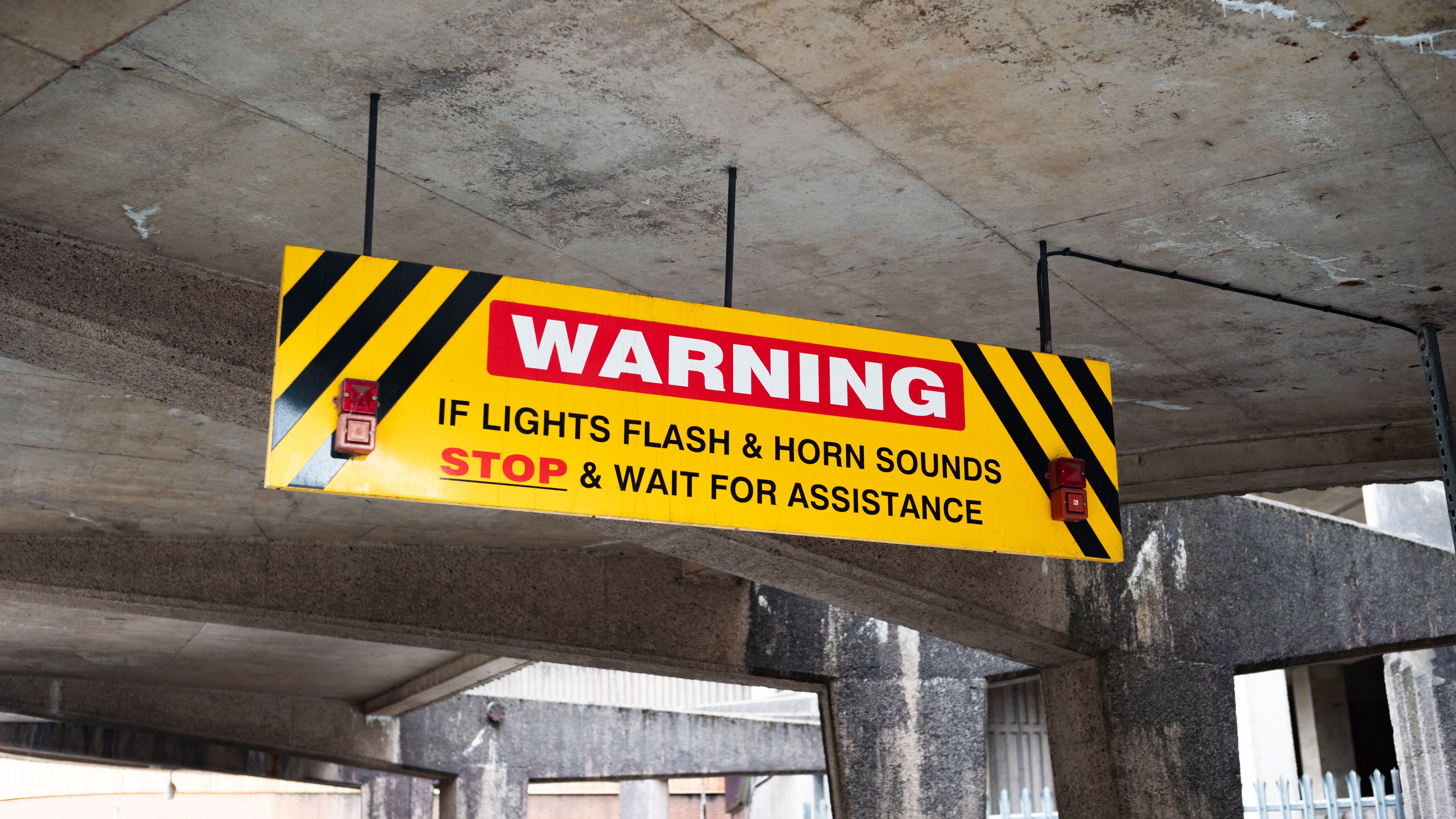 A prominent warning sign in a UK car park advises drivers to stop if lights flash and horn sounds.