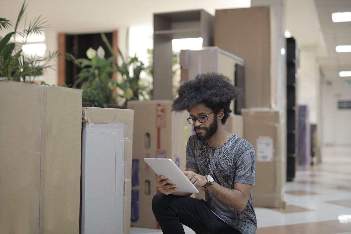 Concentrated young black male employee wearing casual clothes and eyeglasses sitting near boxes and browsing tablet while working in modern workspace