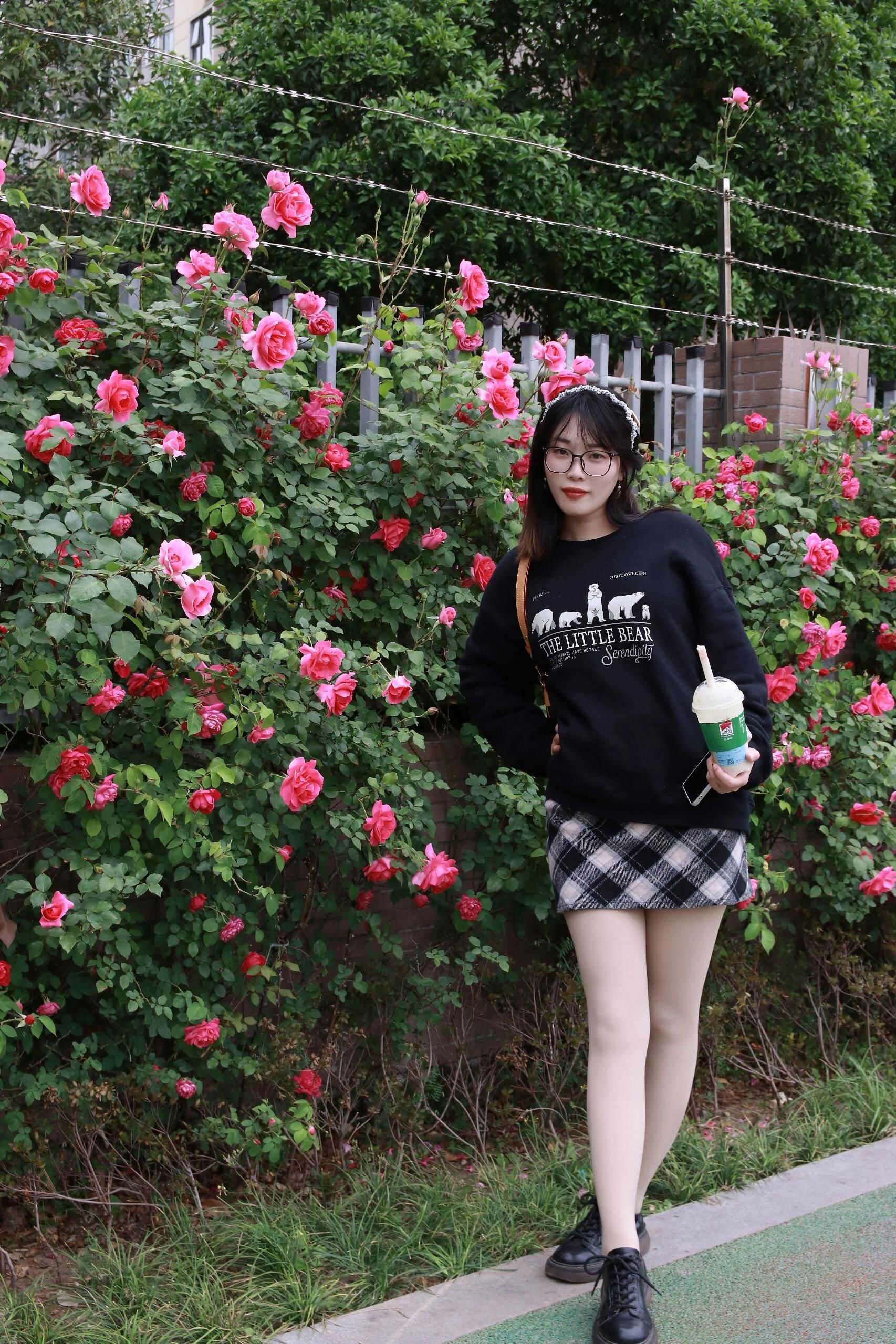 Stylish young woman stands by vibrant pink roses in a Nanjing park, enjoying a refreshing beverage.