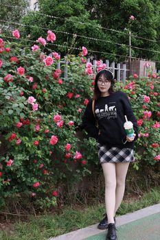 Stylish young woman stands by vibrant pink roses in a Nanjing park, enjoying a refreshing beverage.