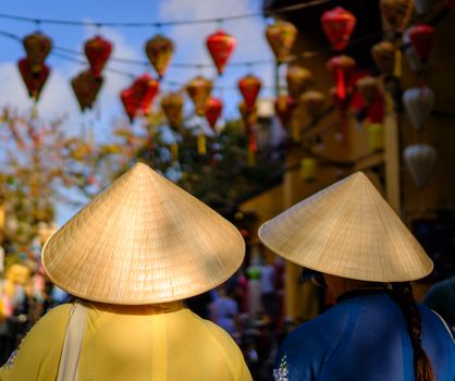 Two people wearing conical hats in vibrant Hội An with colorful lanterns overhead.