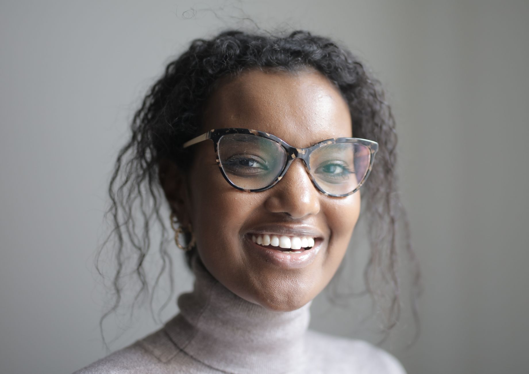 Young African American female in casual clothes and eyeglasses smiling and looking at camera while standing near blurred grey background