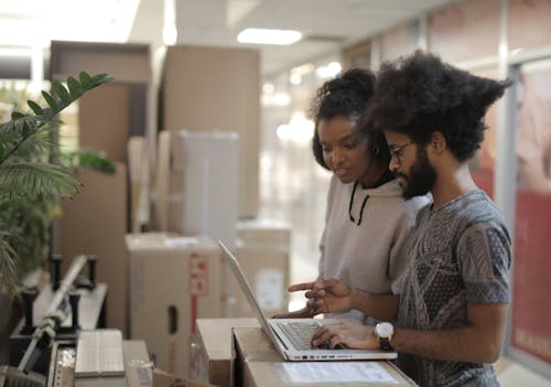 Side view of young African American female and ethnic man wearing casual clothes working on project using laptop while standing near boxes