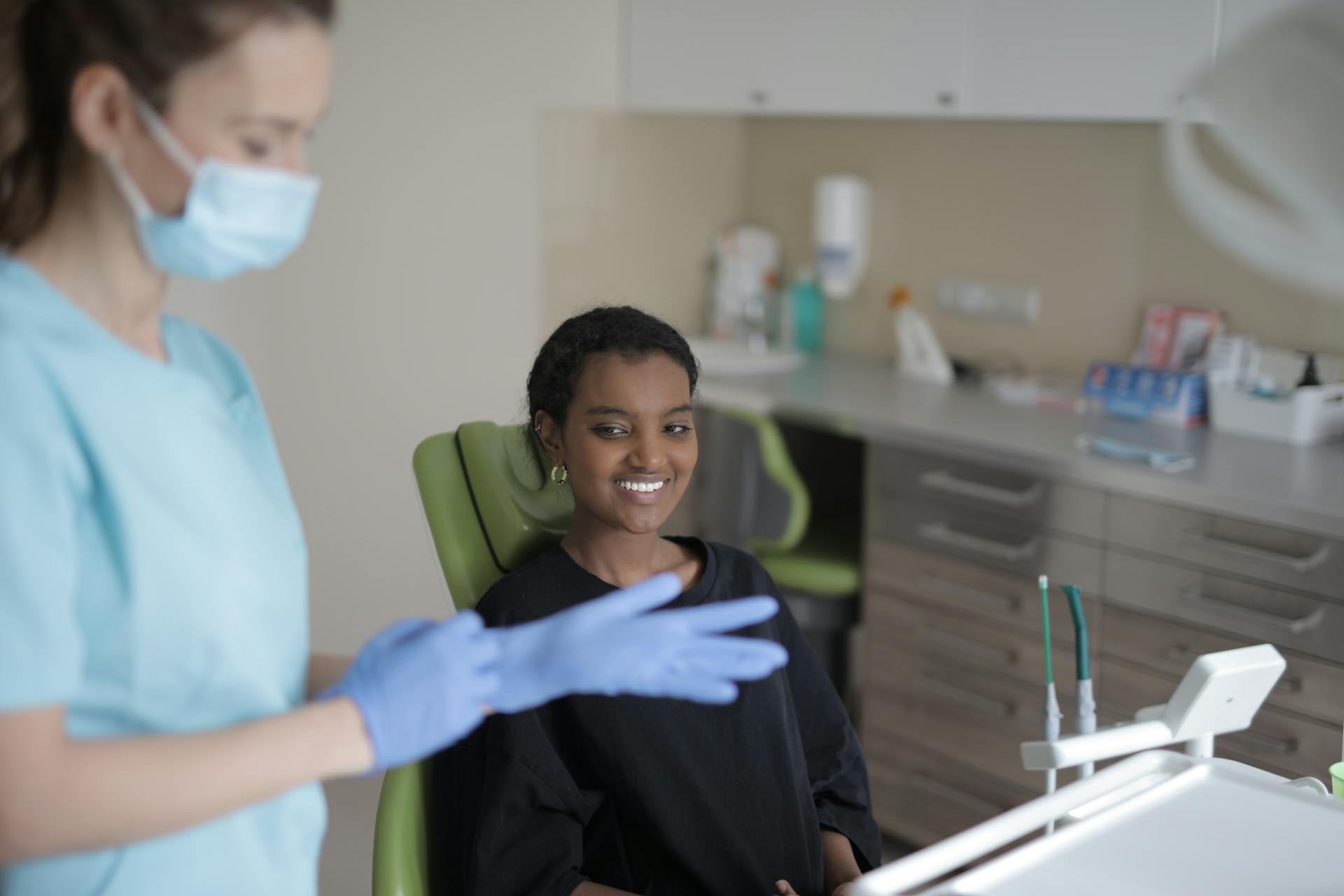 Positive young ethnic female patient smiling while sitting in dental chair and looking at dentist before treatment in modern clinic