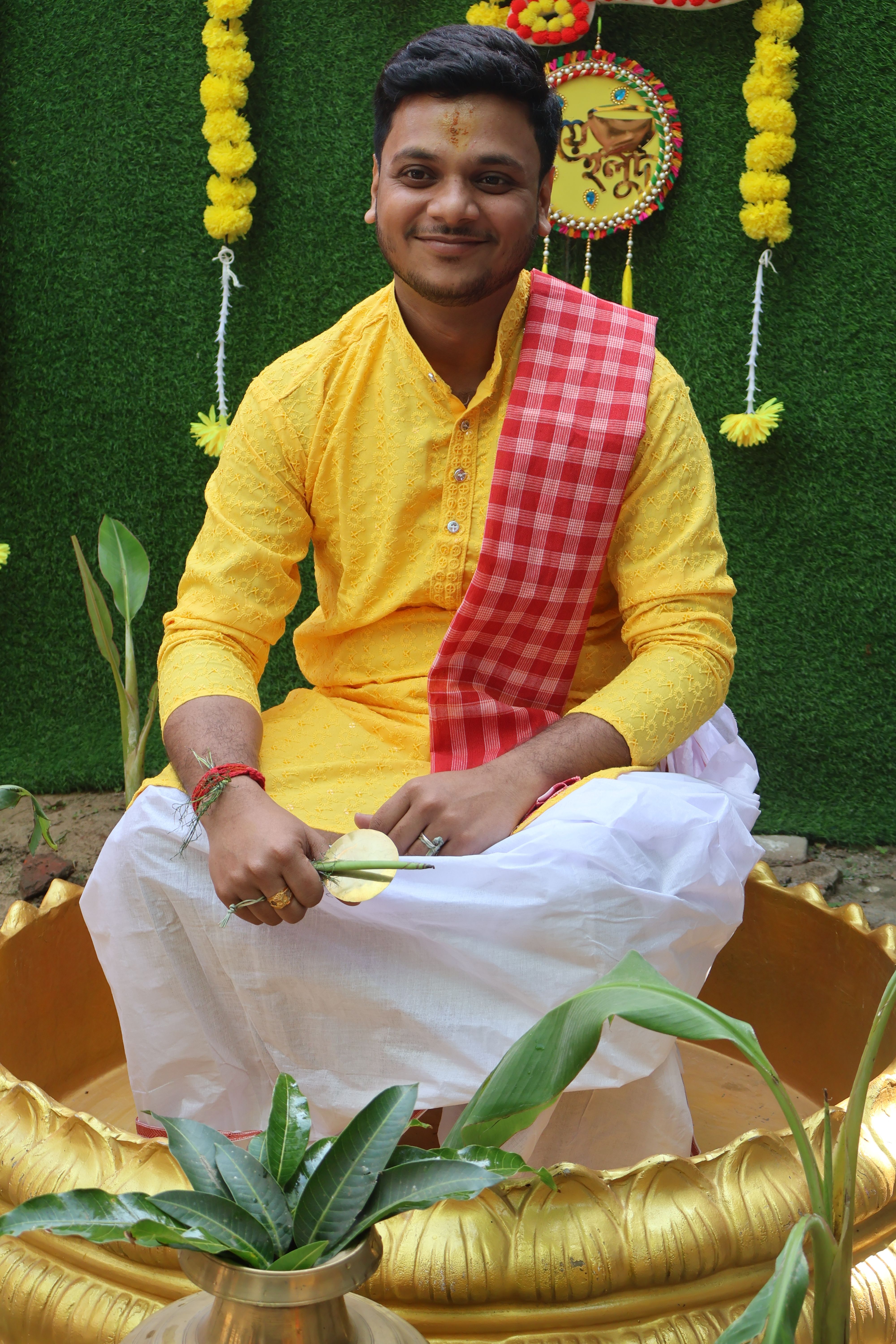 Man in traditional yellow attire sitting joyfully, surrounded by green leaves and festive decor.