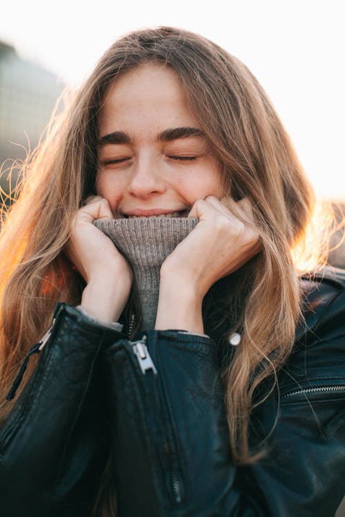Cheerful young woman with eyes closed covering mouth with turtleneck on ...