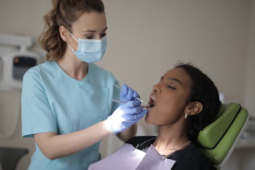 Side view of young female dentist in mask and gloves wearing blue uniform holding medical tools while treating teeth of young ethnic lady in modern clinic