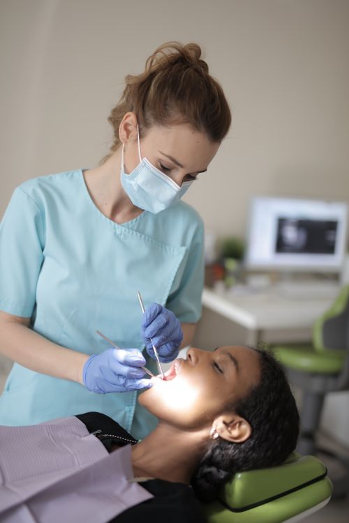 Free Side view of young female dentist in medical mask and latex gloves wearing blue uniform holding medical instruments while treating teeth of female ethnic patient in modern clinic Stock Photo