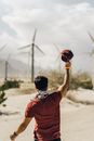 Back view of faceless young guy in casual outfit holding cap in raised hand while standing against wind turbines on sunny day