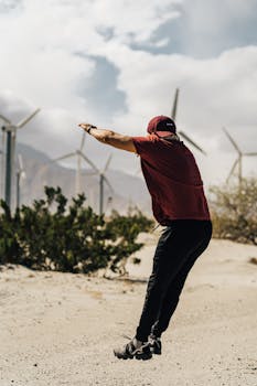 一名身着运动服的年轻运动员在农村风车场旁进行户外锻炼，侧视图中他正在做跳跃深蹲