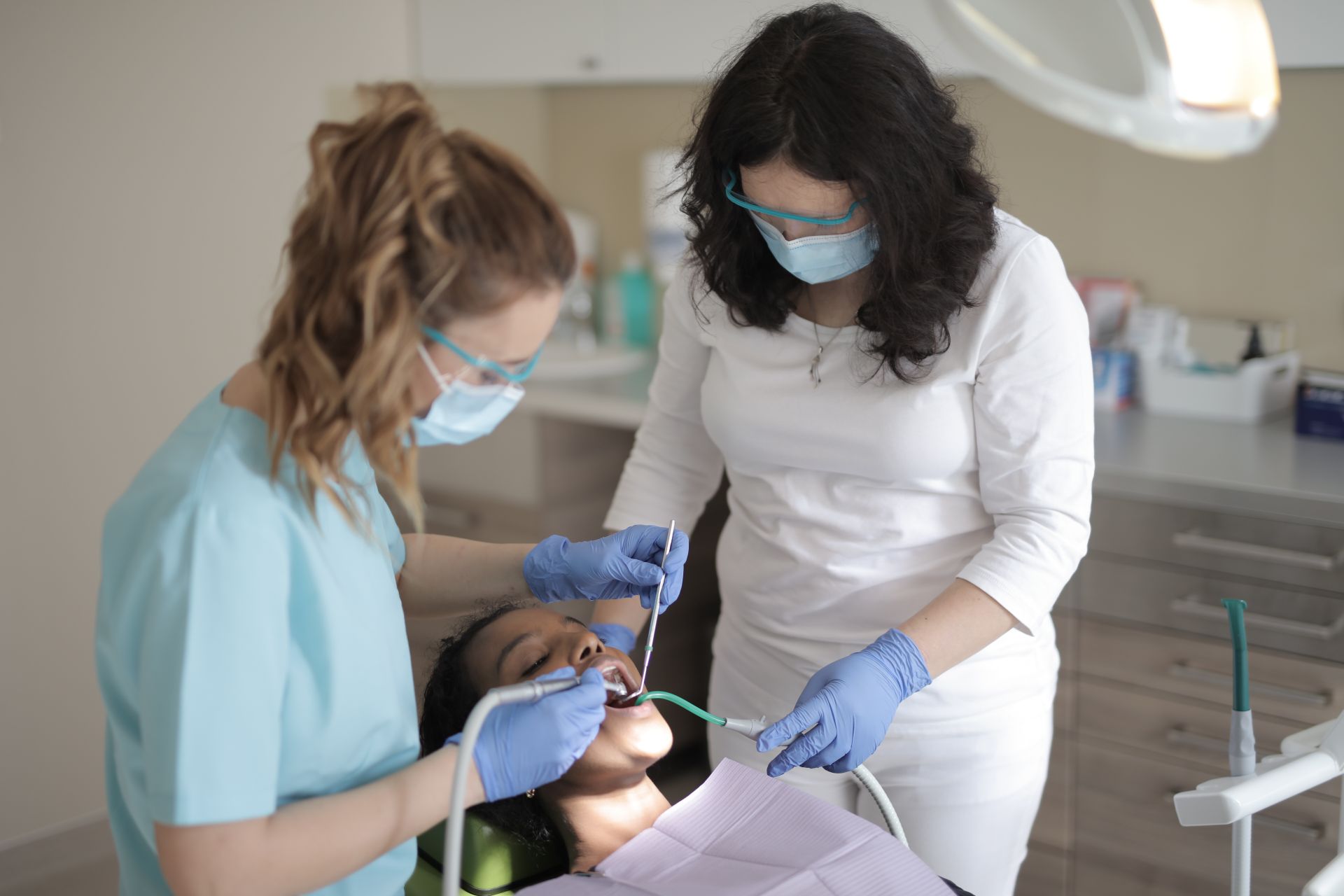 Professional female dentists in medical masks and latex gloves wearing uniform treating teeth with instruments of patient in chair while working in modern clinic