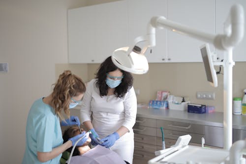 Young female dentist in medical mask and latex gloves curing teeth of patient with assistant using medical instruments in modern dental clinic