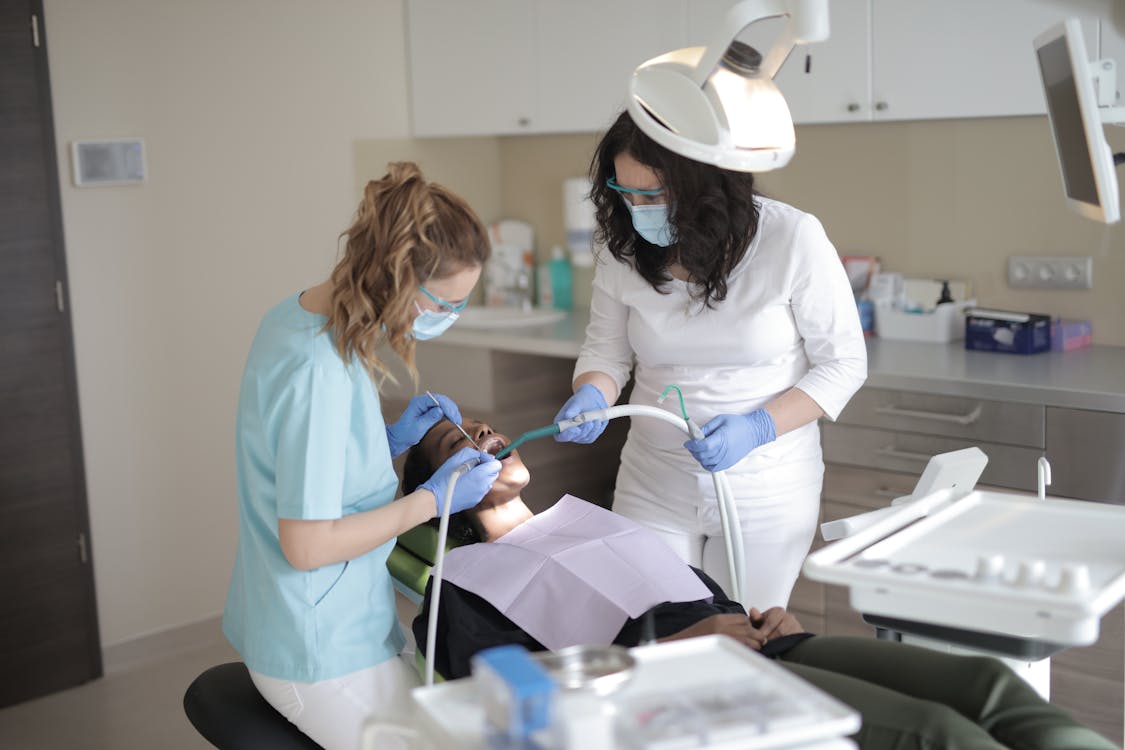 Female dentists in medical masks and latex gloves wearing uniform using medical instruments while treating teeth of female patient in modern dental clinic
