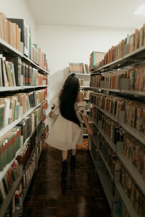 Free A woman in a white dress walks through a library aisle, surrounded by bookshelves. Stock Photo