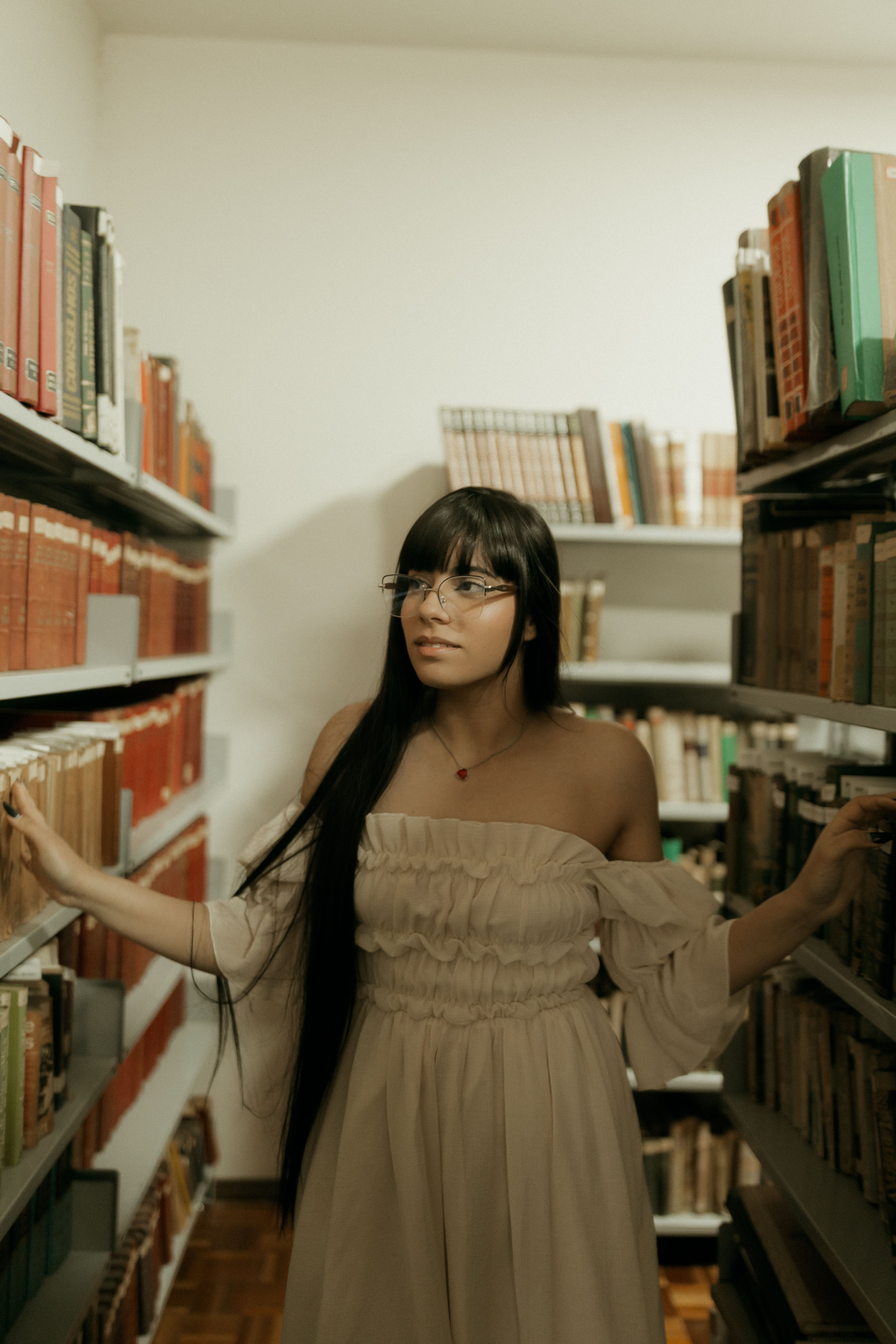 Young woman exploring a library aisle, surrounded by bookshelves. Elegant, studious setting.