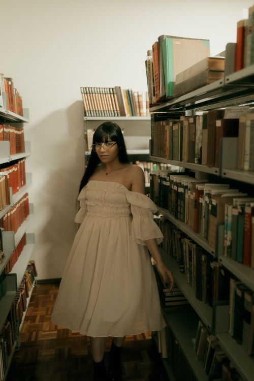 Free A woman in a beige dress walks through library aisles filled with books. Stock Photo