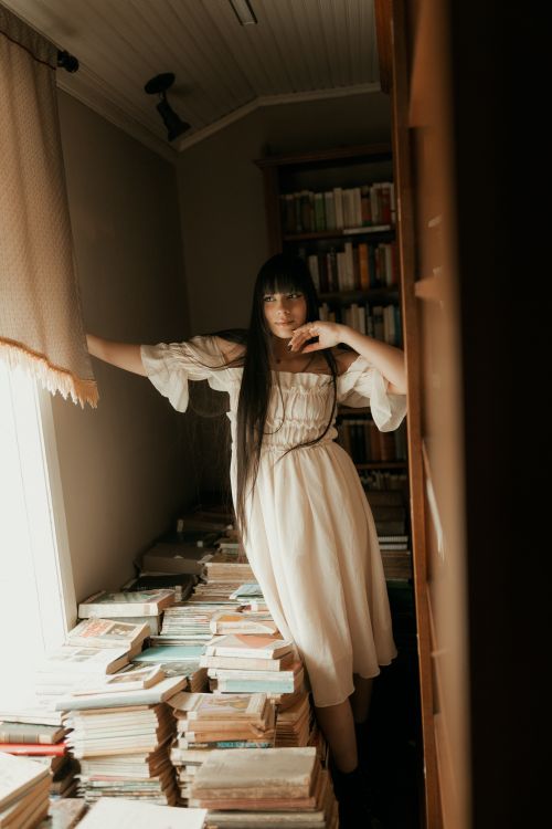 Free A serene image of a woman in a white dress standing amidst piles of books in a warmly lit room. Stock Photo