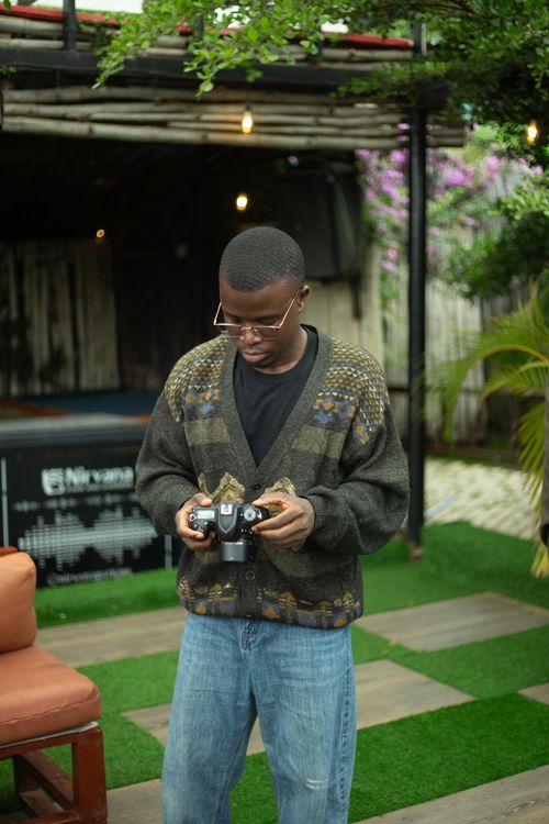Free Young adult holding a camera outdoors in a lush urban setting, surrounded by greenery. Stock Photo