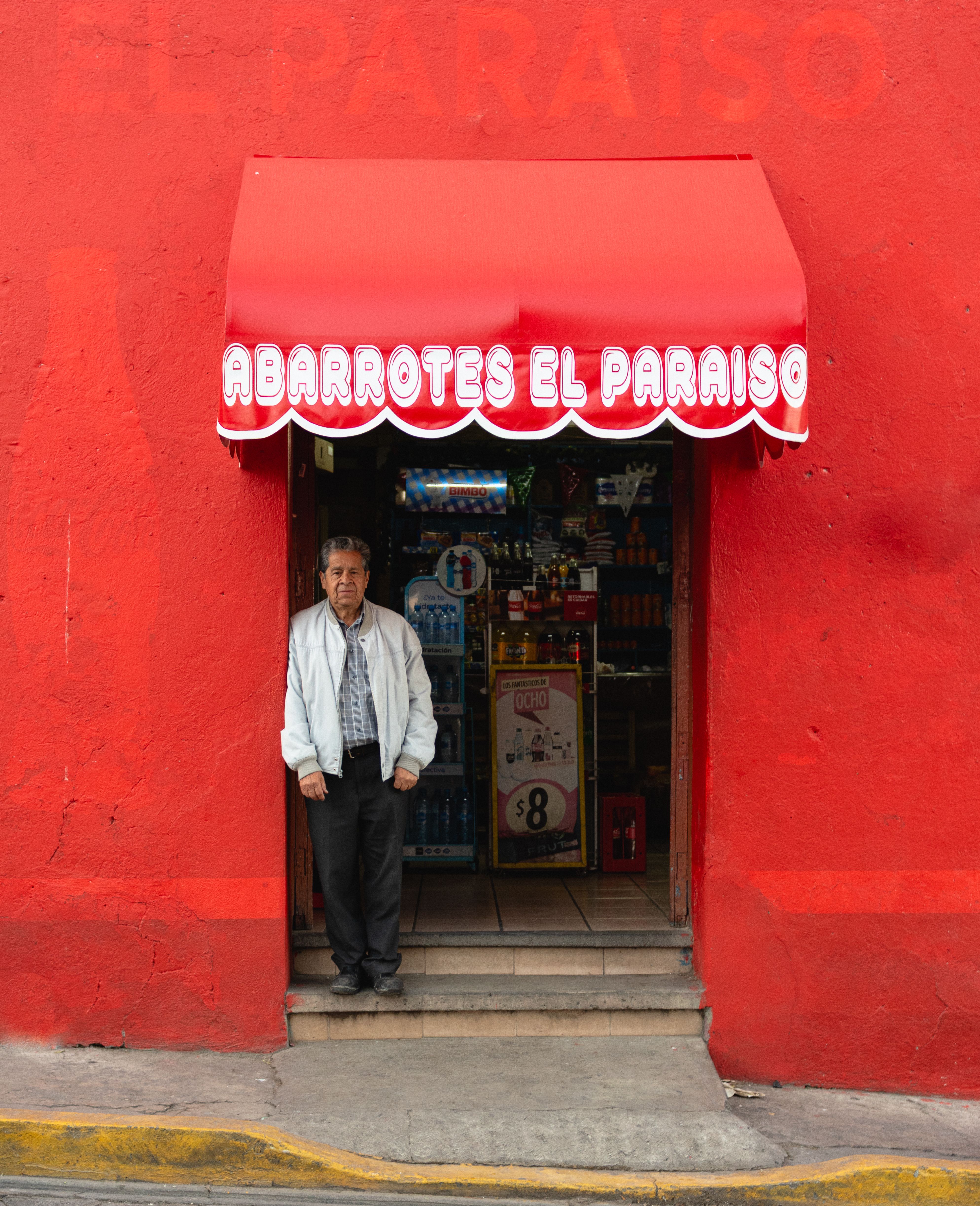 Senior man stands at the entrance of a vibrant red grocery store in a city.