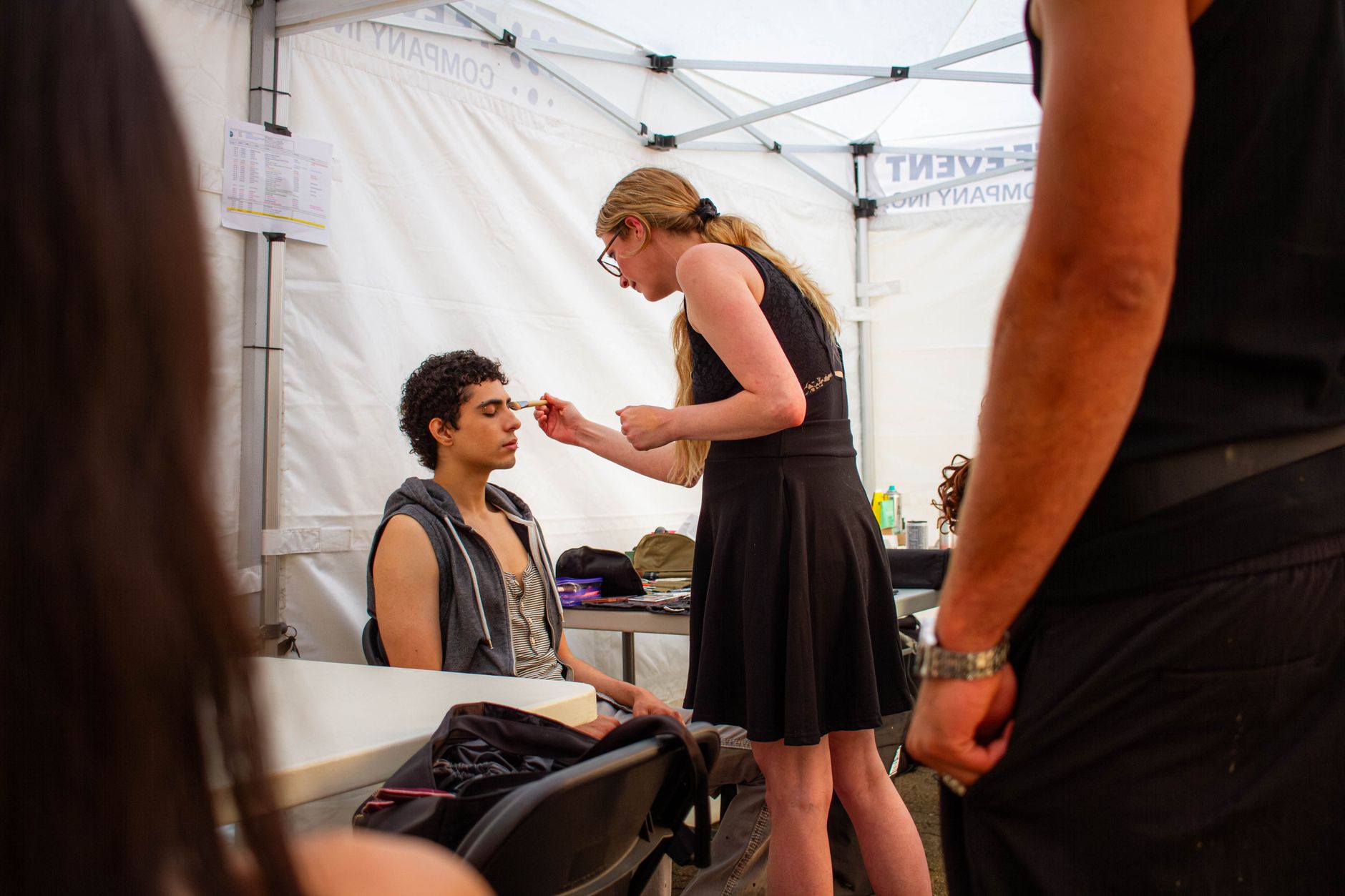 Makeup artist applying cosmetics to a young man in an event tent setup.