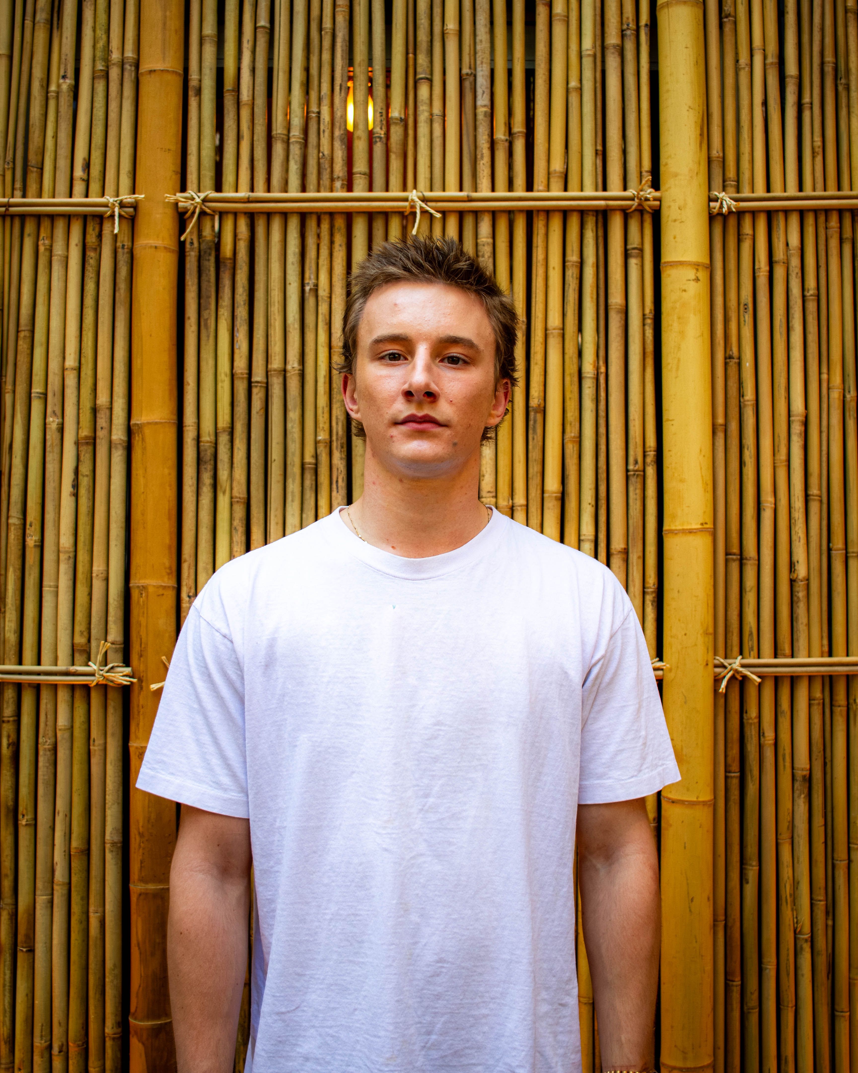 A young man wearing a white shirt stands confidently against a bamboo backdrop, showcasing a blend of modern fashion and natural elements.