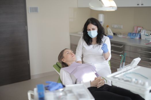 Female dentist in medical mask and latex gloves wearing uniform while sitting near chair with patient and explaining details of treatment while working in modern clinic