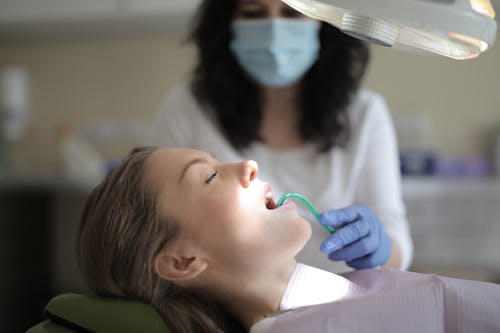 Side view of female dentist in medical mask and latex gloves using saliva ejector while treating teeth of woman sitting in chair in modern dental room in clinic