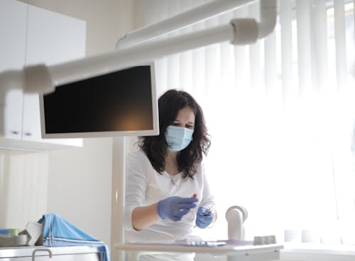 Free Serious female dentist in medical mask and latex gloves wearing white uniform preparing instruments for treatment in light modern dental room in clinic Stock Photo