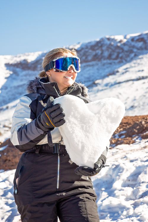 Free A woman in winter attire holds a heart-shaped snow in a snowy mountain landscape, conveying warmth. Stock Photo