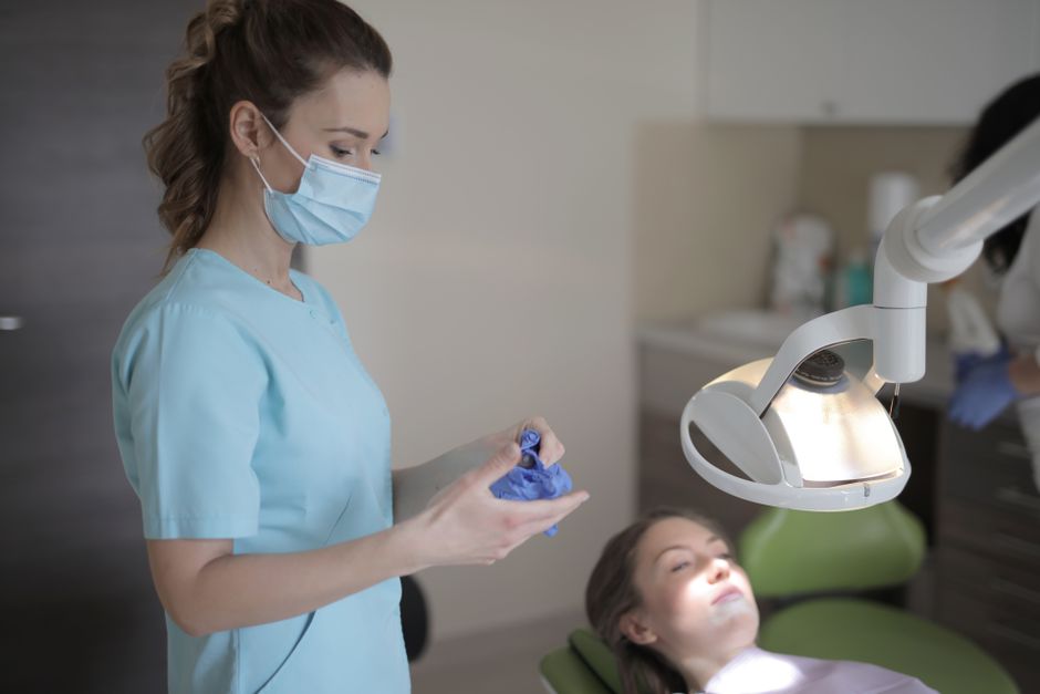 nighttime oral health - Side view of young female dentist in medical mask and uniform taking off latex gloves after treatment patient in modern dental room in clinic
