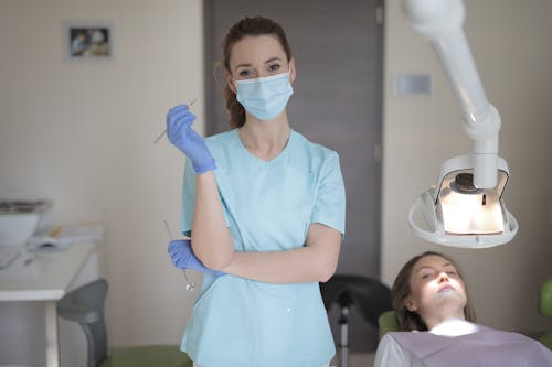 Young female dentist in medical mask and latex gloves wearing blue uniform standing with instruments in hands and looking at camera while working with patient in modern dental room