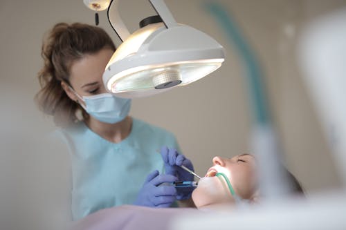 Side view of young female dentist in medical mask and latex gloves wearing uniform using professional equipment while curing teeth of patient in modern dental room in clinic