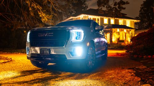 Free A white GMC Yukon XL SUV with headlights on, parked in front of a lit house at night. Stock Photo
