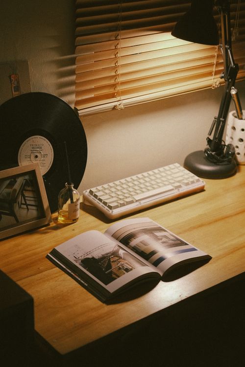 Free Warm, cozy workspace featuring a keyboard, open book, desk lamp, vinyl record, and wooden blinds. Stock Photo