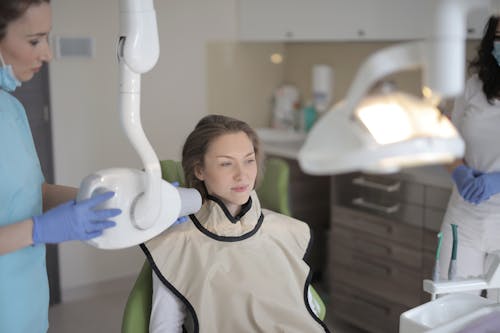 Young female dentist in latex gloves and uniform using modern medical equipment while working with woman sitting in dental chair in clinic