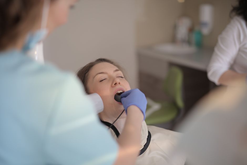 Young female dentist in latex gloves and blue uniform checking teeth of young patient during medical procedure in modern dental room in hospital