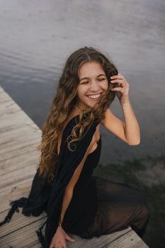 Smiling woman with long hair sitting on a wooden pier by the water.