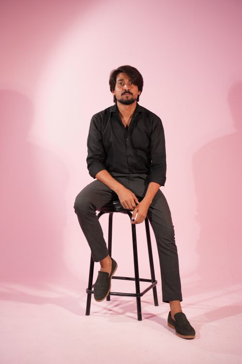 Free Confident man in black attire sitting on a stool, posed against a soft pink background. Stock Photo
