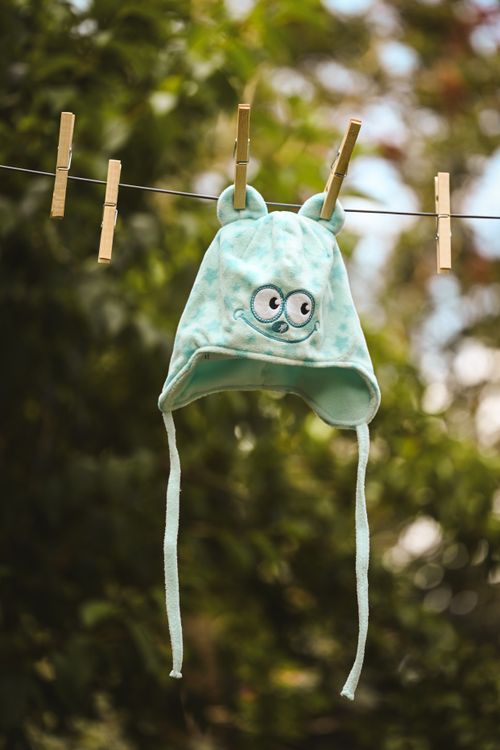 Free Adorable blue baby hat with eyes design hanging on a clothesline with clothespins against a leafy background. Stock Photo