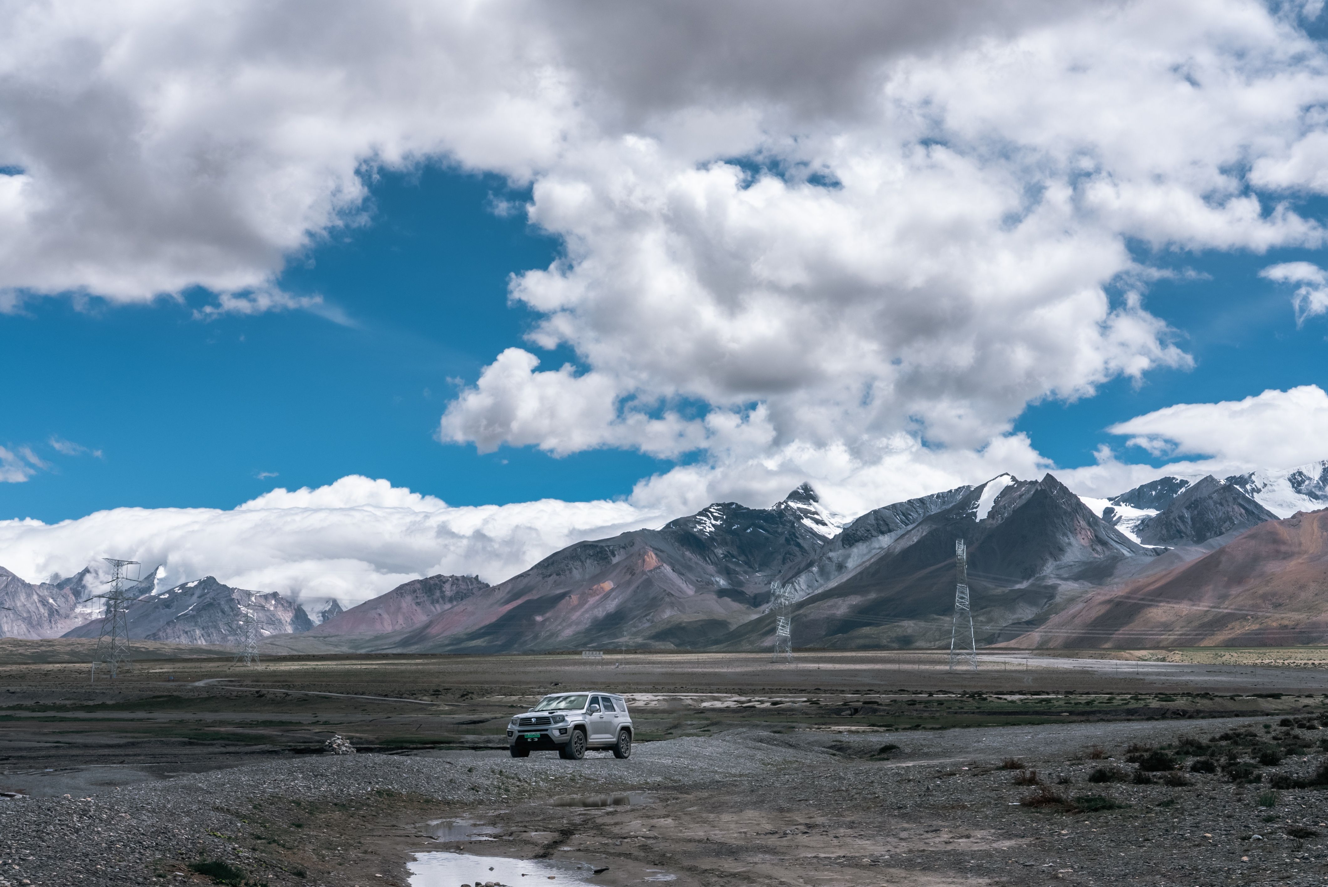 A lone SUV travels through a vast, rugged mountainous terrain under a dramatic sky.