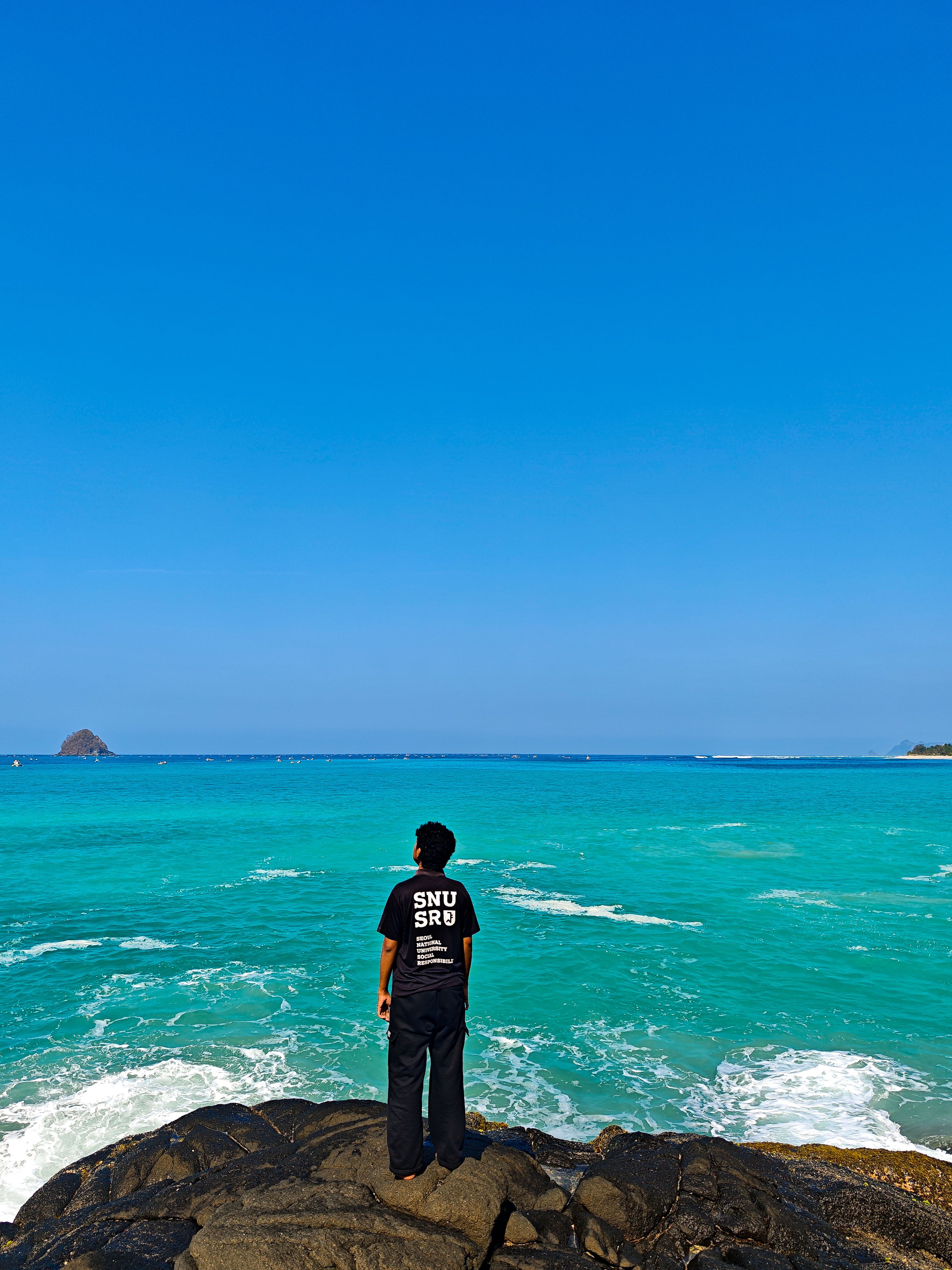 A person stands on rocks, gazing at the turquoise sea under a clear blue sky in West Nusa Tenggara, Indonesia.