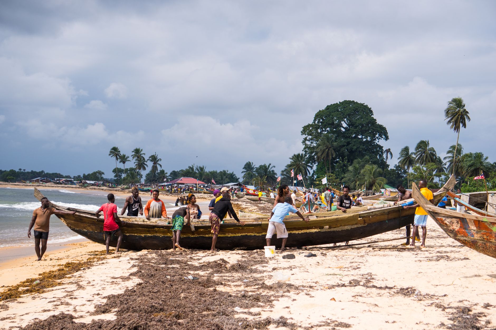 Locals collaborate to pull a canoe onto the sandy shores of a Liberian beach on a cloudy day.