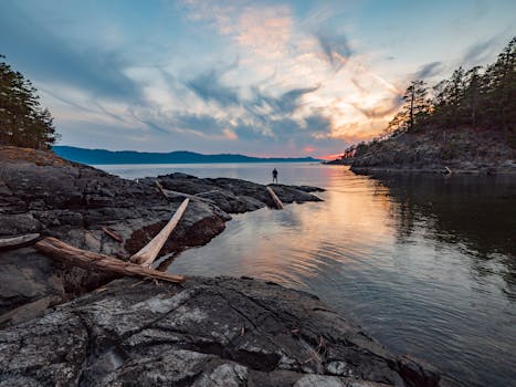 在加拿大不列颠哥伦比亚省的一个湖边，一个人站在岩石岸边，独木舟旁，捕捉宁静的日落