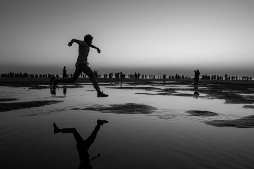 Free A person captured in silhouette jumps over a reflective shoreline during twilight, with a crowd in the background. Stock Photo