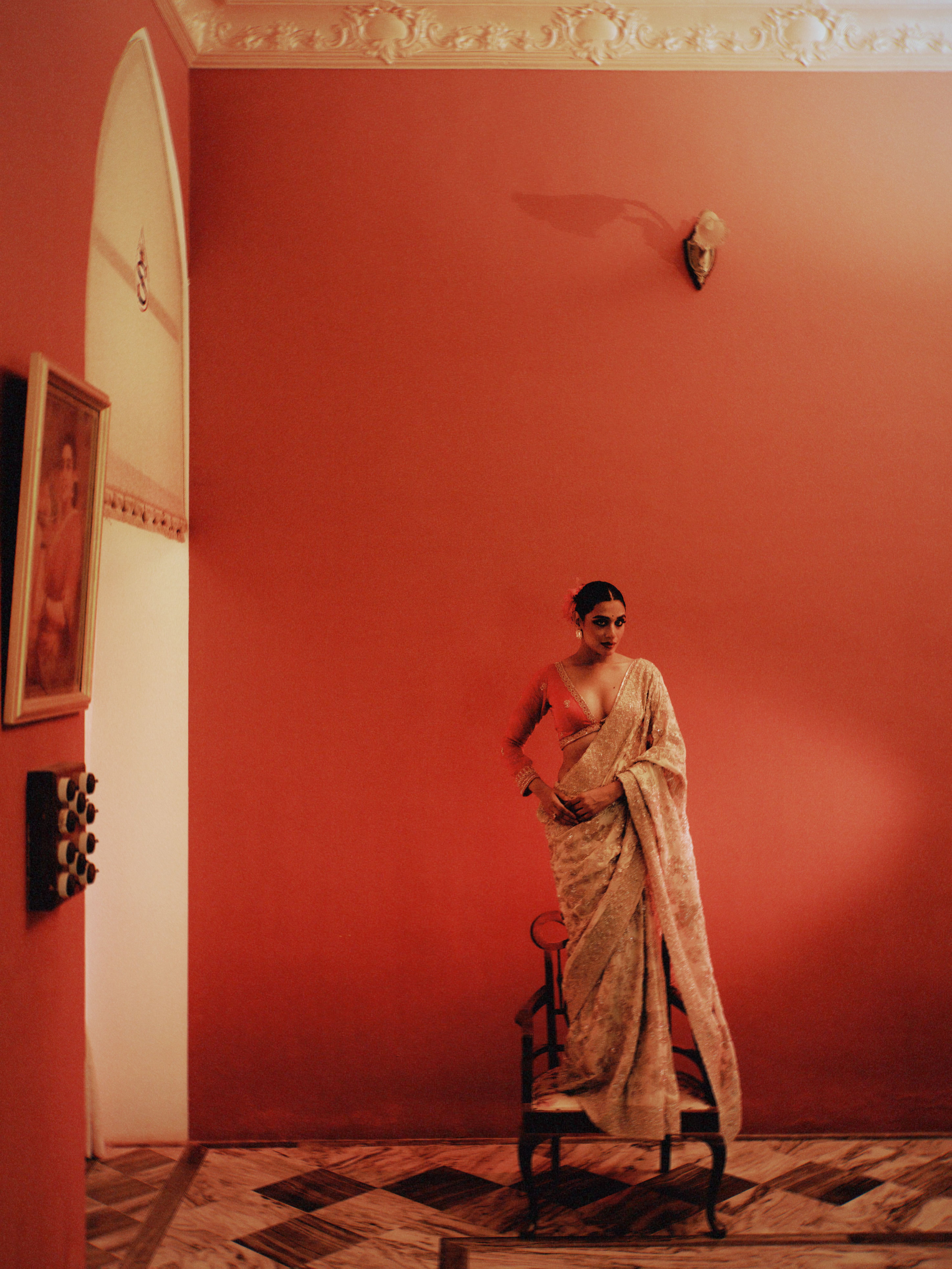 Woman in saree posing in richly colored, indoor setting with vintage decor.
